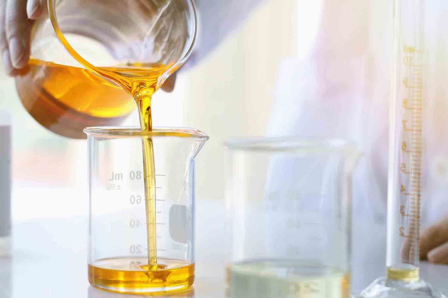 Golden oil being poured from a glass beaker into a laboratory measuring cup with measurement markings visible.