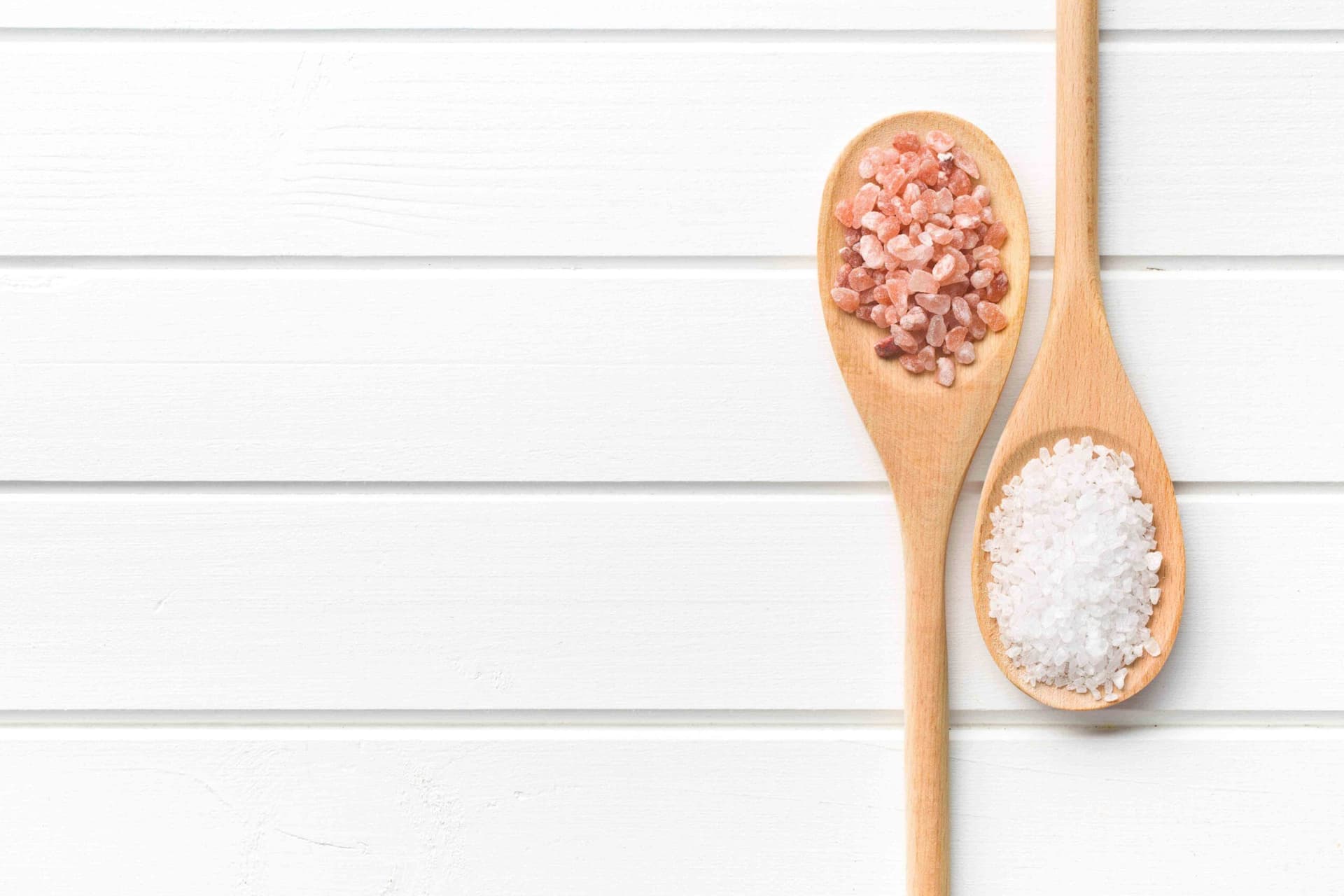Two wooden spoons holding pink Himalayan salt and white sea salt on white wooden background.