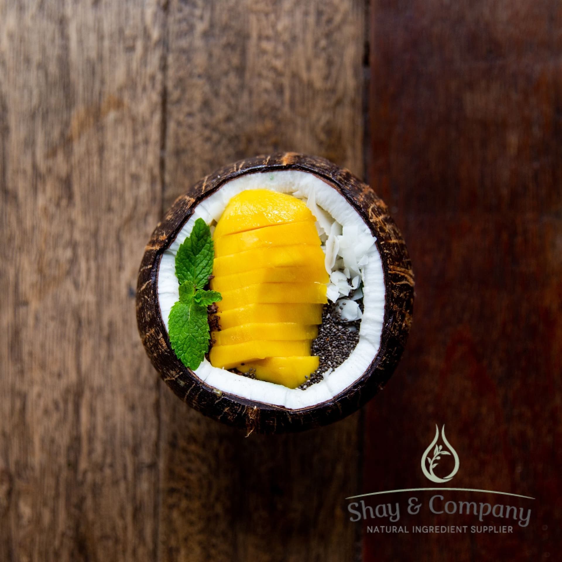 Halved coconut with mango slices, chia seeds, and mint leaf on rustic wood surface by Shay & Company.